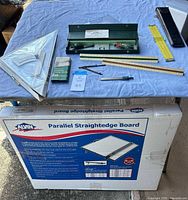 Photo of drafting tools including rulers, slide rule in case, lettering set and drafting pen on a blue cloth with original box of Alvin Parallel Straightedge Board below.
