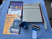 Boston 2612 paper cutter with photo paper packs, black mug, and box of Swingline staples on blue cloth background.