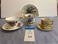 Three teacup and saucer sets including the Regency bone china set with purple flowers and gold rims in center, photographed with white backdrop.