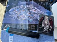 Photo showing Millennium Falcon schematic poster, coffee table book 'Dressing a Galaxy' and black storage tube