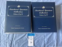 Two closed navy blue binders side by side on fabric backdrop, gold foil text visible reading Volume I of II and Volume II of II