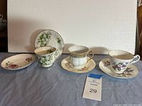 Four teacup and saucer sets displayed in a row against a white background on a blue surface, showing various floral designs and gold trims.