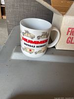 Front side of white ceramic mug showing Hummer M998A2 Series text and vehicle illustrations, placed on plastic surface with open cardboard box in background.