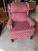 Red patterned upholstered wingback armchair with carved wooden claw-foot front legs, cushioned headrest and seat, and padded arms. Shows minor wear and needs cleaning.
