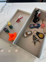 Various vintage toy figures, small vehicles, and keychains laid out in two cardboard boxes on a white surface.