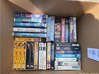 Photo showing 23 assorted VHS tapes with visible titles including Indiana Jones trilogy, Under Siege, Maverick, Apollo 13, Vacation and others.