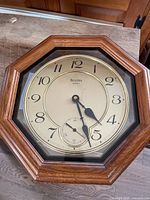Full view of the Bulova wall clock showing the wooden octagonal frame, clear glass face, beige clock dial with black numerals and hands, and a small seconds subdial at the bottom.