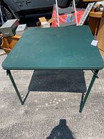 Green folding card table outdoors on pavement with metal legs and green vinyl top, square 33" by 33" surface, 28" tall.