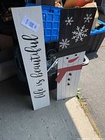 Three decorative wooden boards stacked outdoors; showing the text, snowflakes and snowman designs
