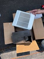 Nutone bathroom exhaust fan grille being held above fan housing inside cardboard box on outdoor surface.