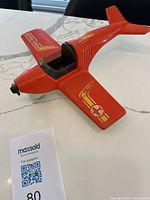 Top angled view of red plastic Fisher-Price toy airplane showing cockpit and decals, placed on white marble surface.