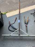 Full overview of three garden tools: large metal rake, hedge shears, and hand scythe laid out on concrete ground.