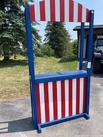 Front view of the handcrafted kids play stand painted blue with red and white vertical stripes, showing the window opening and countertop shelf.