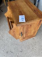 Wooden side table with magazine rack on one side and closed cabinet door on the other side, showing natural wood grain and metal handle.