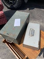Photo showing two metal boxes: a larger grayish-green metal file box with a handle on the front, and a smaller light gray cash box with a latch and handle on top.