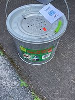 Photo showing the galvanized steel floating minnow bucket with lid and handle with a green grip placed on ground.