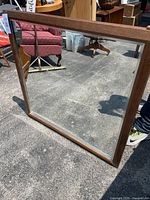 Full view of the large square mirror with visible wood frame reflecting an outdoor scene including a red chair and person's shadow.