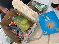 Box containing assorted office supplies including file folders, envelopes, markers, pens, binder clips, Post-it notes and small stationery items.