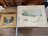 Two framed winter landscape artworks displayed side by side on a wooden table, showing overall condition and framing