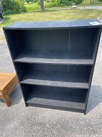 Front view of the black particle board shelf showing three shelves and signs of wear.
