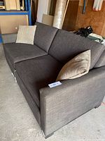 Grey brown fabric sofa bed with two beige throw pillows, shown from angled front view in a garage with concrete floor and various items in background.