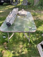 Full view of rectangular outdoor table with folded beige umbrella and round plastic stand on top, placed on grass outdoors.