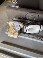 Photo of beige rotary dial telephone and white electric iron on a gray surface next to a couch.