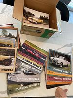 Multiple 'Turning Wheels' magazines laid out next to a cardboard box filled with more magazines showing classic Studebaker cars on covers.