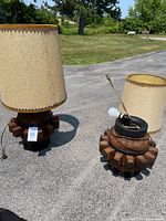 Two 1970s wagon wheel style table lamps, one with shade attached and one with shade removed, positioned outdoors on pavement with grass background