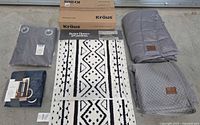 Overall photo showing all items included in lot: Pendleton weighted blanket and cover folded, two curtains in gray and navy, boxed Kraus faucet, Better Homes & Gardens bath rug set in black and white geometric pattern.