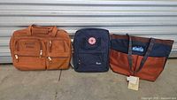 Lot showing the three bags from left to right: Absolut Vodka computer bag, Chick-Fil-A backpack, and Kavu tote bag on concrete floor in front of metal storage unit.