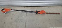 Full view of the Black+Decker cordless pole hedge trimmer laid flat on concrete, showing whole length including handle, shaft and trimming blade.