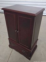 Closed two-door wood jewelry armoire with bottom drawer and dark finish.