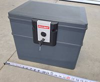 Gray First Alert fireproof lock box with key lock and keys, placed on ground with measuring tape showing approximately 16 inches in width.