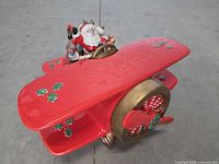 Side angled view showing Santa figure piloting the red biplane with holly decorations and gold accents on propeller and engine.