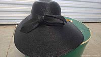 Black woven hat with black ribbon bow and decorative pin on the brim, positioned on top of the green original storage box.
