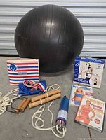 Photo showing a large black exercise ball, jump rope, resistance band with clip, vintage Apollo Exerciser and exercise manual