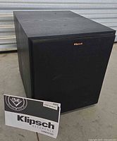 Three images showing a black Klipsch R-100SW powered subwoofer with a black grille and the Klipsch logo on the front. The subwoofer has a textured finish and sits on the floor. A Klipsch Reference Subwoofers user manual is placed in front in all images.