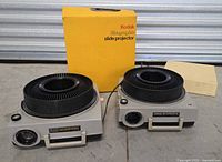 Two Kodak Ektagraphic Slide Projectors Model E-2 positioned side by side with original yellow Kodak box and operating manual behind them on a flat surface.