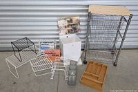 Full lot photo showing the assortment of organizers including a metal wire rolling cart with wooden top, white plastic bins, wine glass racks, silverware trays, risers, and pegboard hooks on a concrete floor.