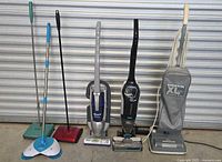Photo showing 6 floor cleaning tools including 3 upright vacuum cleaners, 1 stick vacuum, and 3 manual sweepers. All items arranged side by side against a metal garage door.