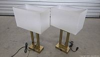 Pair of modern table lamps with white rectangular fabric shades and brass-tone metal bases, cords visible, shown from front angle.