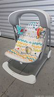 Front view of baby swing showing multi-color chevron patterned fabric, overhead bar, soft animal toys, and white base.