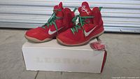 Front and top view of red and green Nike LeBron 9 basketball shoes with original box showing LeBron branding and extra laces beside shoes.