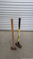 Pair of two sledgehammers, one with wooden handle, one with black/yellow composite handle, standing upright on concrete floor in front of roll-up garage door.