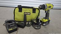 Photo showing Ryobi cordless drill, two 18V batteries, battery charger, side handle attachment, and carrying bag.