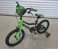 Full view of green and black Schwinn 16 inch Piston bike with training wheels attached, positioned outdoors on concrete.