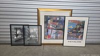 Four framed pieces standing outdoors against a shutter door: two black and white photographs, one abstract art print in a gold frame, and one colorful Sausalito print