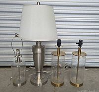 Four modern/contemporary style table lamps on concrete floor with shutter background. Two brass and glass lamps are cylindrical and matching, one silver and glass lamp is cylindrical with silver base, and one brushed silver formal style lamp with white shade.