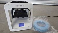 Front view of the white Toybox 3D printer and spool of blue filament wrapped in plastic, showing the compact frame and components inside.